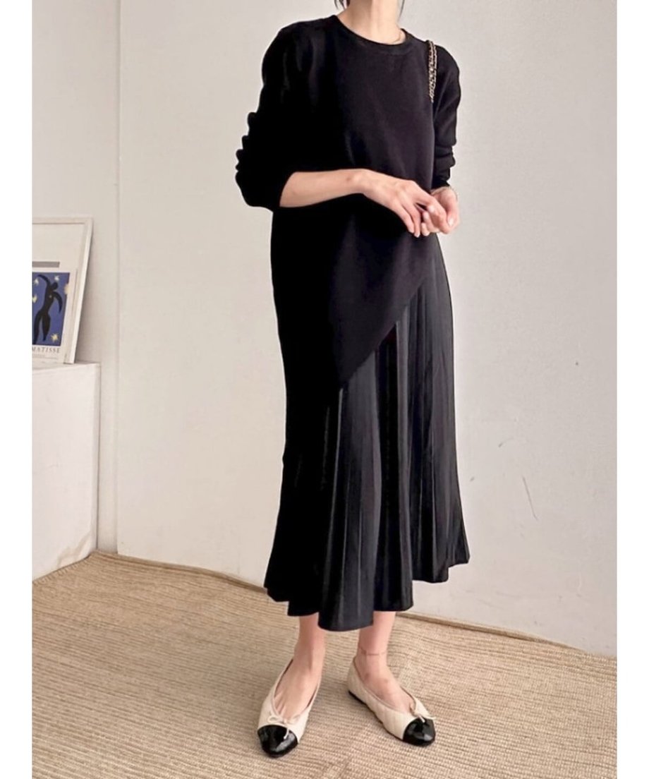 ブラックのアシンメトリーニットとプリーツスカートセットアップをを着た女性が立っているシーン。