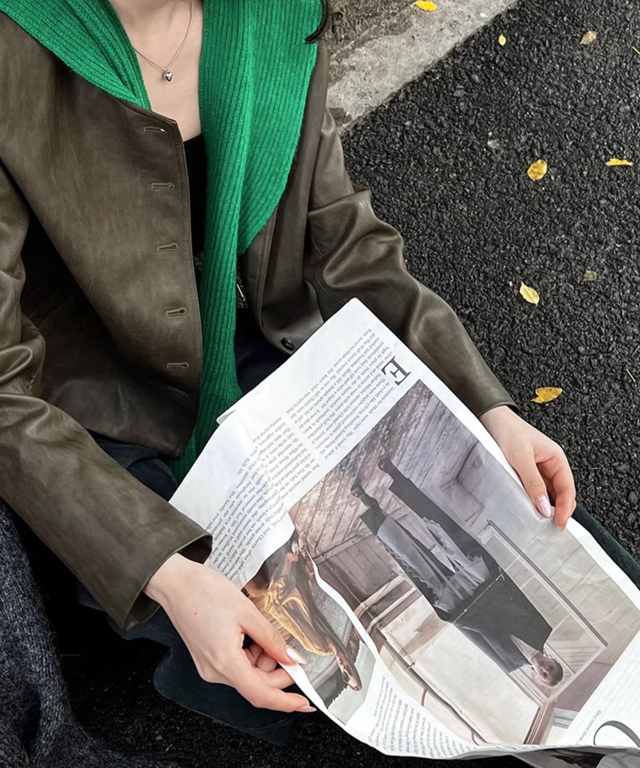 カーキのフェイクレザージャケットに、緑色のニットを肩掛けした女性のクローズアップ。新聞を持っている。