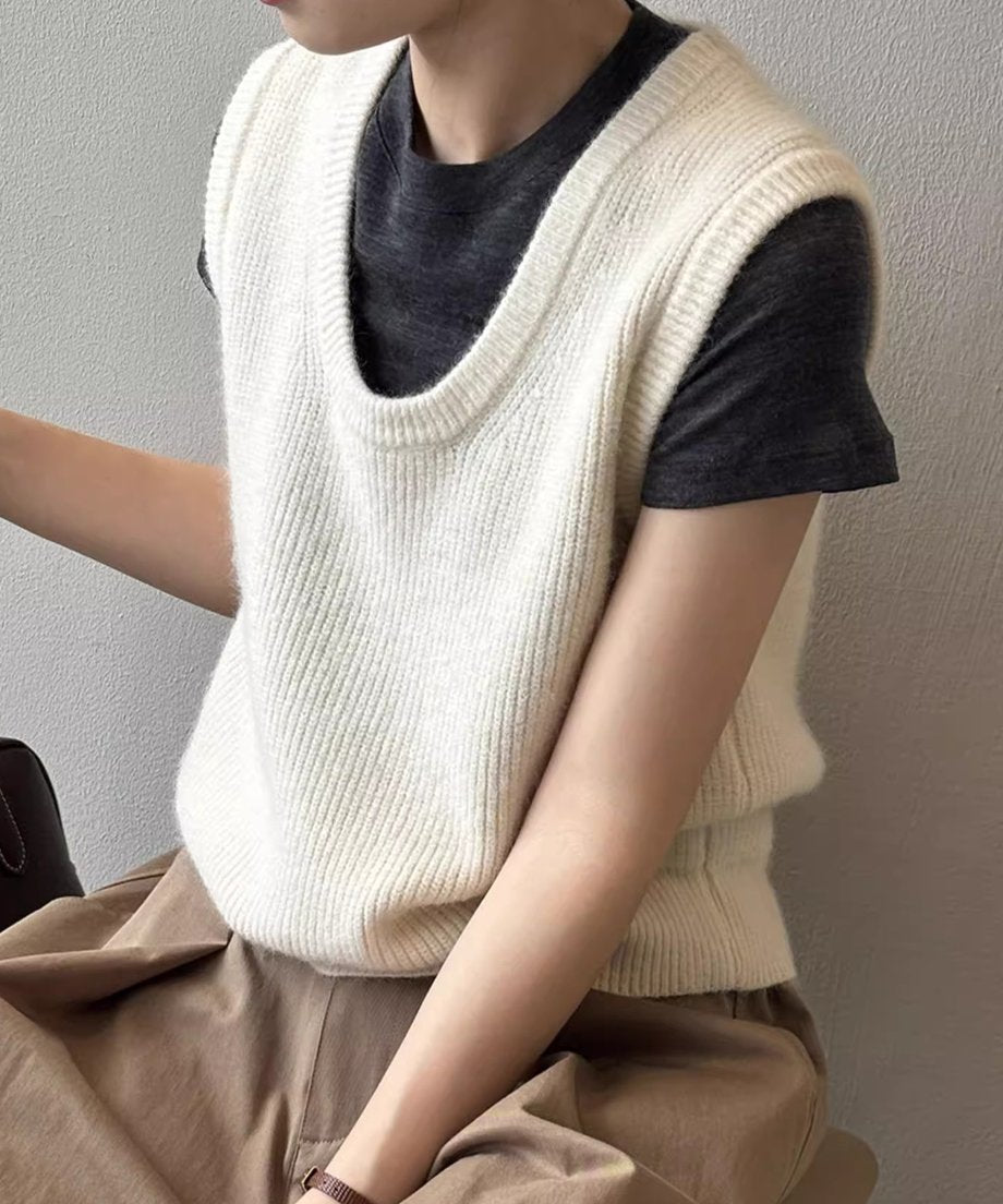 オフホワイトのUネックニットベストを濃いグレーのTシャツと茶色のパンツにレイヤードし着用している女性のクローズアップ。