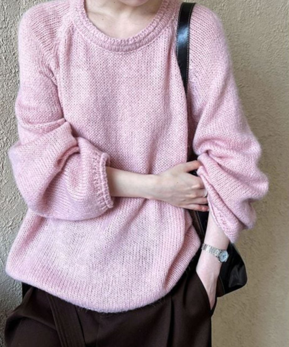 ピンクのローゲージニットを着用し、右肩に黒いバッグをかけた女性の、バストアップ写真。時計をつけた手首が見える。