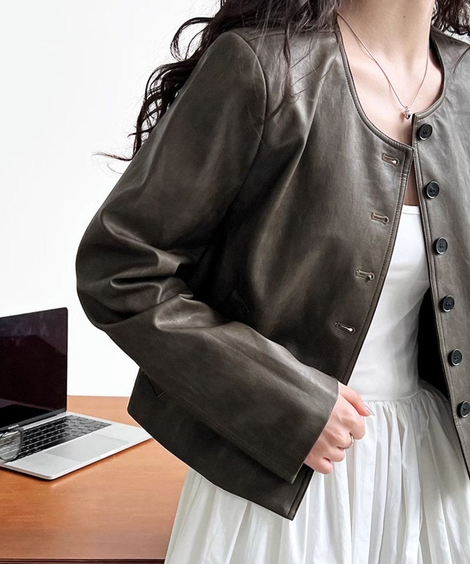カーキのフェイクレザージャケットを羽織った女性のクローズアップ。白のノースリーブワンピースと合わせており、手前にはノートパソコンが置かれたデスクがある。