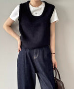 ブラックのUネックニットベストを白Tシャツと濃い色のデニムにレイヤードして着用している女性の全身像。首元にはシルバーのネックレス、左手にバッグが見える。