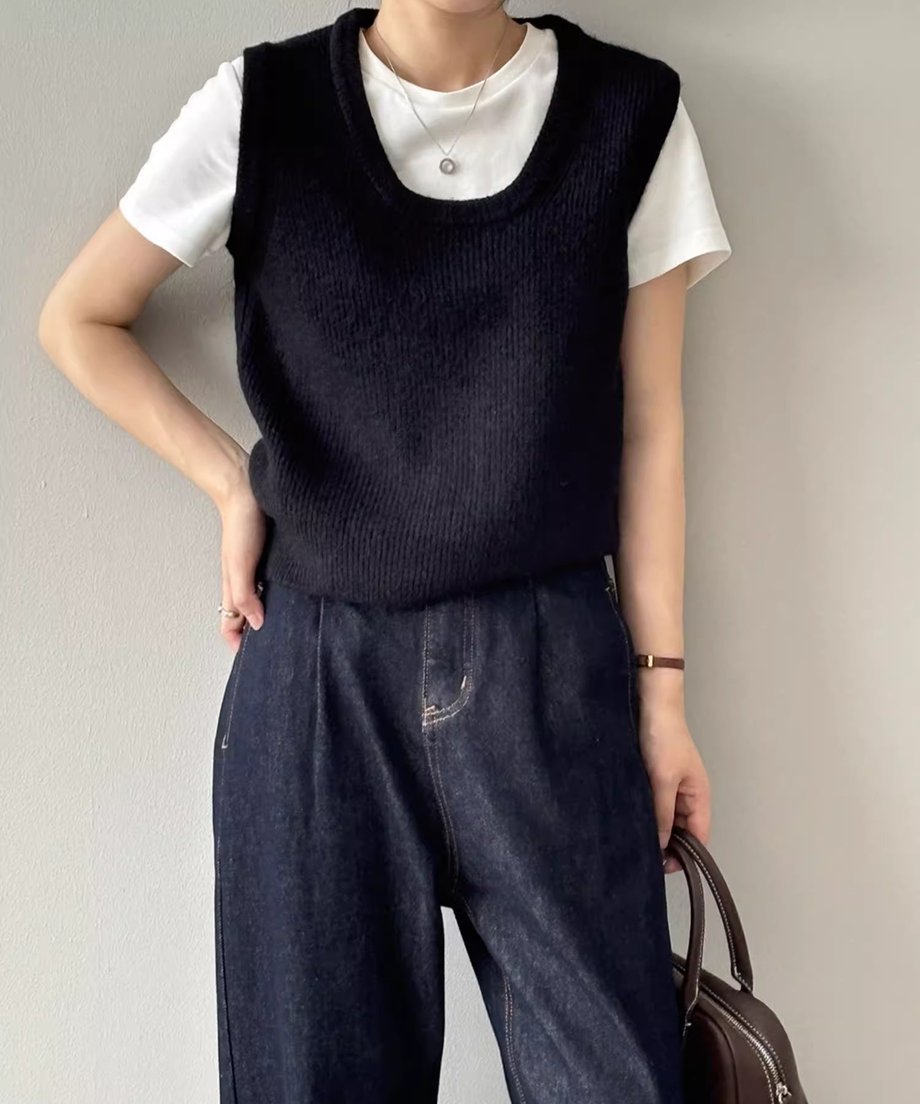 ブラックのUネックニットベストを白Tシャツと濃い色のデニムにレイヤードして着用している女性の全身像。首元にはシルバーのネックレス、左手にバッグが見える。