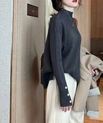 ダークグリーンのモックネックニットを着用し、薄い色のワイドパンツを履いた女性の側面写真。袖口に3つのボタン付きスリットが見える。