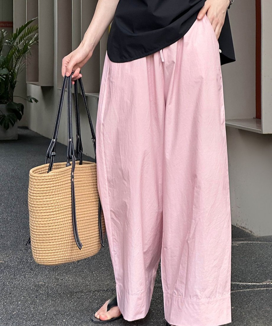 ピンクのワイドレッグパンツと黒のトップスを着用し、ナチュラルな編みバッグを持った、横からのクローズアップ写真