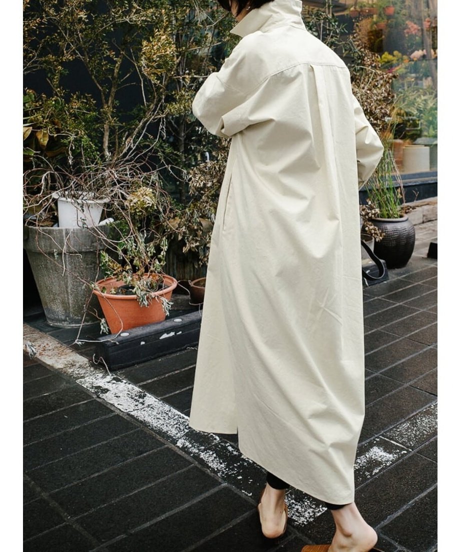 アイボリーのロングシャツワンピースを着用した女性の後ろ姿。屋外の植栽を背景に、裾が長く広がる様子。