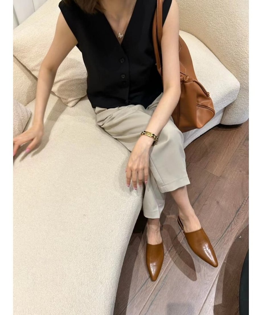 黒いノースリーブトップスとベージュのクロップド丈パンツ、ブラウンのポインテッドトゥミュールを着用し、ソファに座る女性。茶色の大きなバッグを横に置いている。