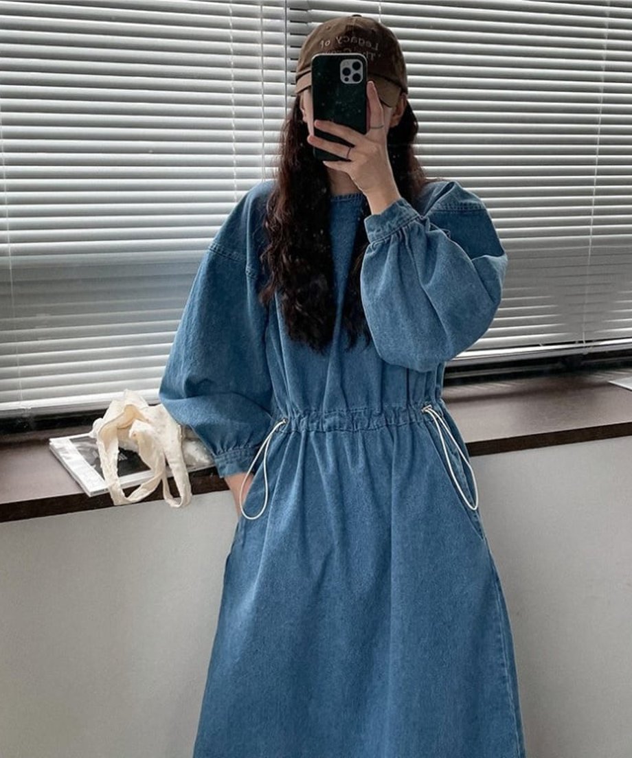 青のデニムドローストワンピースを着て、茶色のキャップをかぶった女性が、ブラインドの前でスマートフォンで自撮りをしている様子。