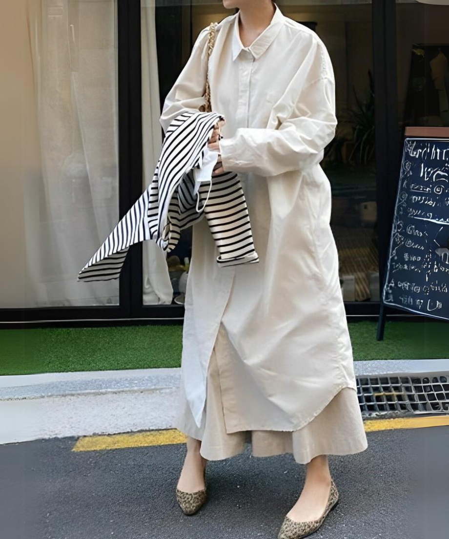 ロングシャツワンピースを着用し、ベージュのキルティングバッグを持ち、ボーダー柄のトップスを抱えて屋外を歩く女性。