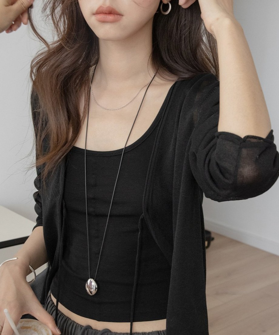 ブラックのドロストシアーシアーカーディガンと黒のタンクトップを着用した女性のバストアップ。片手に飲み物を持ち、耳に手を添えている、シンプルな夏の重ね着スタイル。