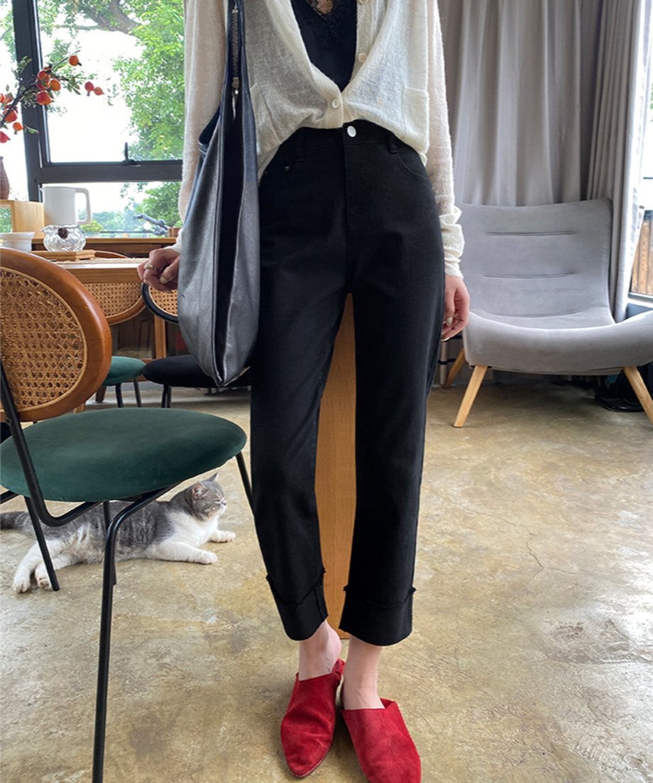 屋内のカフェ風の場所で、黒の裾カフス付きストレートパンツを着用した女性の全身。白いカーディガンと赤のスエード調ミュールを合わせており、足元には猫がいる。