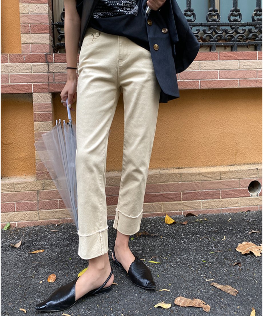 壁を背景に、アプリコットの裾カフス付きストレートパンツを着用し、黒のジャケットとTシャツを合わせた女性。黒のポインテッドトゥミュールを履き、透明な傘を持っている。