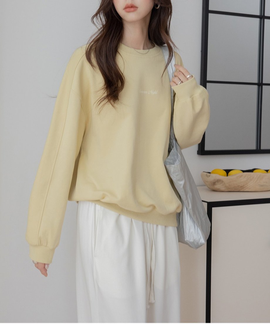 イエローのオーバーサイズスウェットと白のワイドパンツを合わせた全身コーディネート。女性は淡い色のショルダーバッグを持ち、室内で立っている。