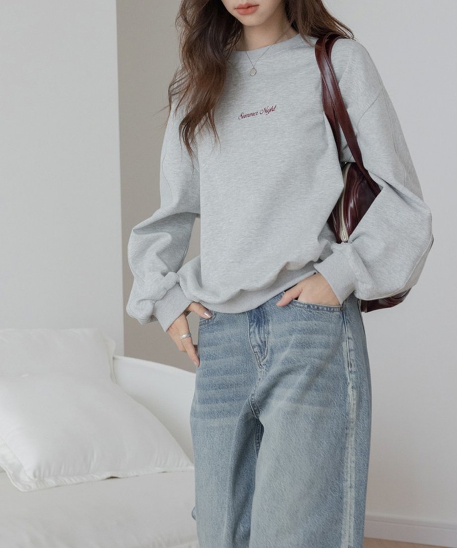 グレーのオーバーサイズスウェットを着用した女性。胸元には「Summer Night」の赤茶色の刺繍ロゴがあり、左手にシルバーのリングとバングルを着用し、茶色のバッグを肩にかけている。