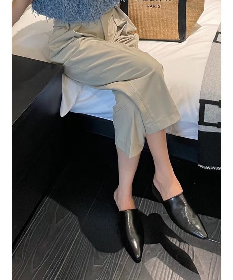 ベージュのワイドパンツを着用し、ベッドの端に座っている女性の足元。ブラックのポインテッドトゥミュールを履いている