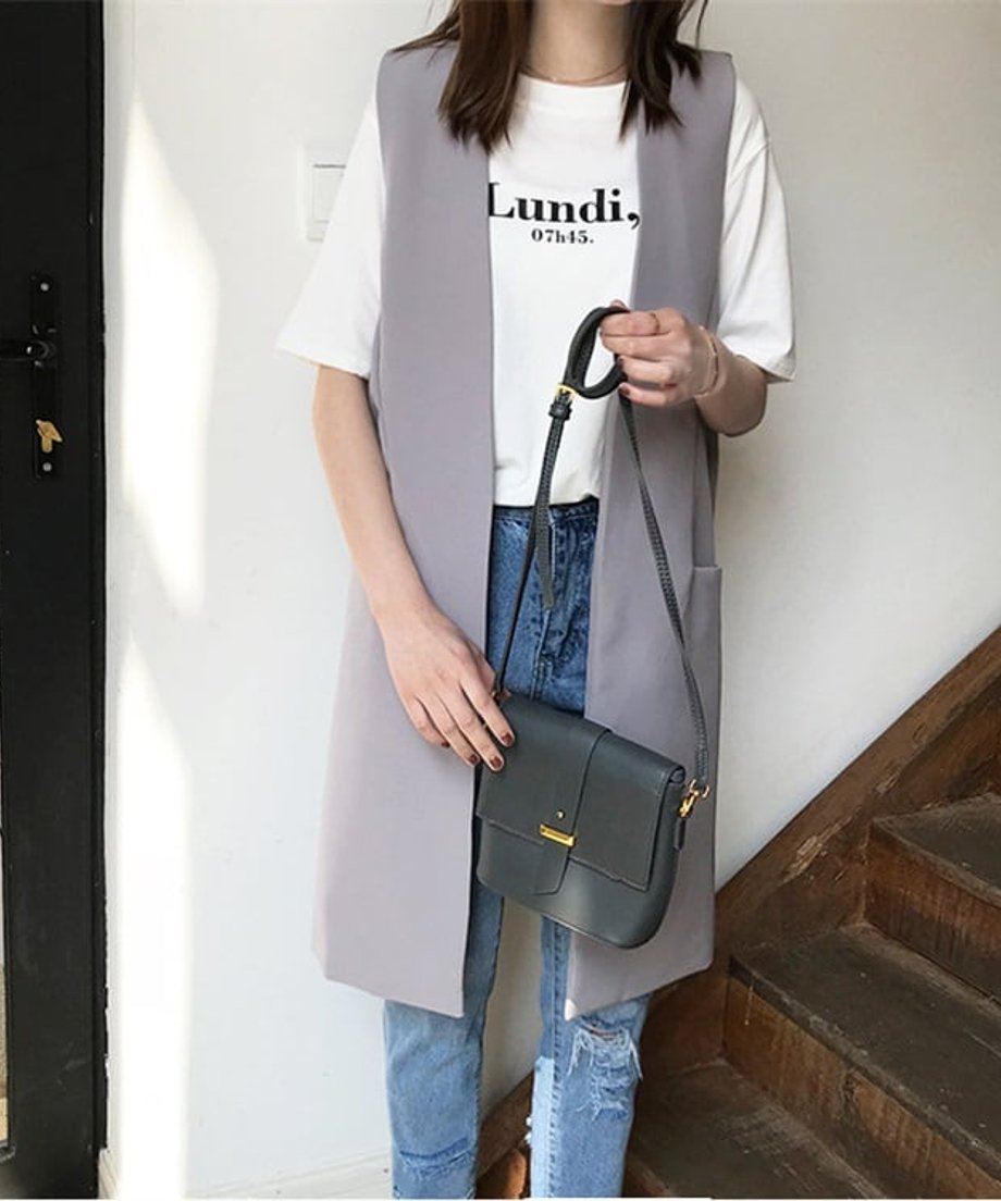 白いロゴTシャツとデニムにライトグレーのロングベストを着用した女性が、ショルダーバッグを両手で持っている。