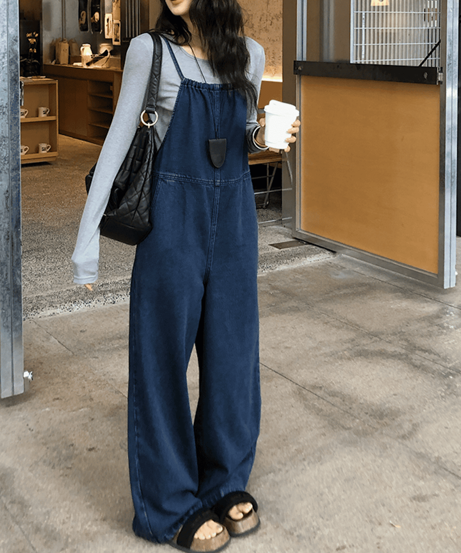ダークブルーのデニムサロペットを着た女性が、カフェの前でコーヒーを持って立っている。