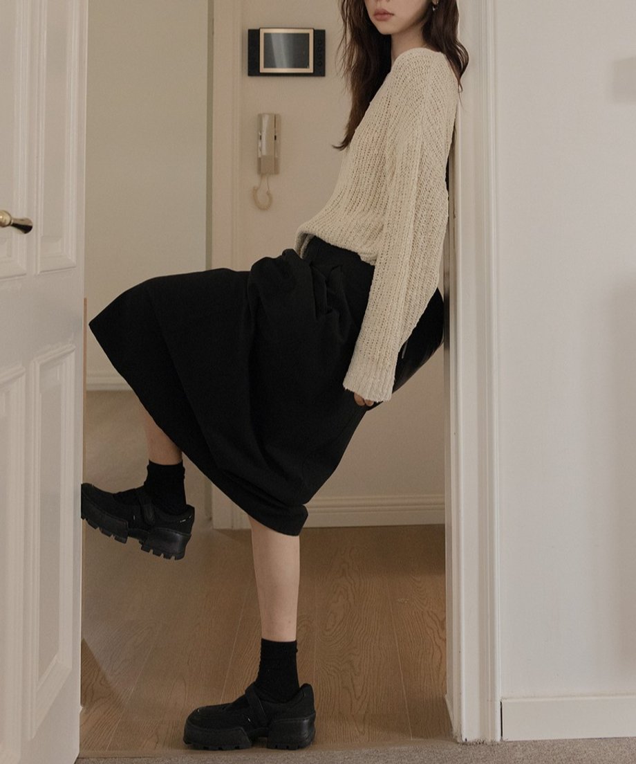 ベージュのニットと黒のプリーツスカートを着用し、壁にもたれかかる女性。ボリュームのあるシューズがコーディネートを引き立てる。