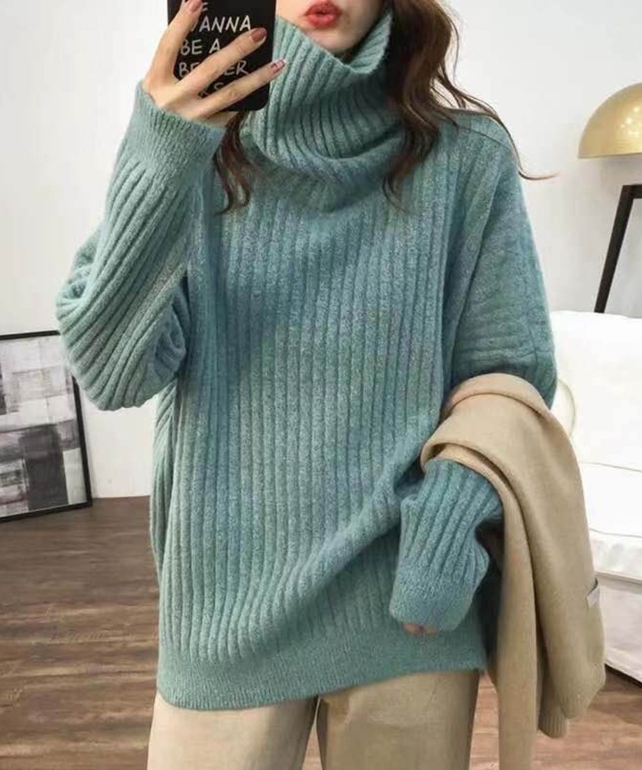 グリーンのオーバーサイズタートルネックニットを着用し、ベージュのストールを腕に抱えた女性の上半身。