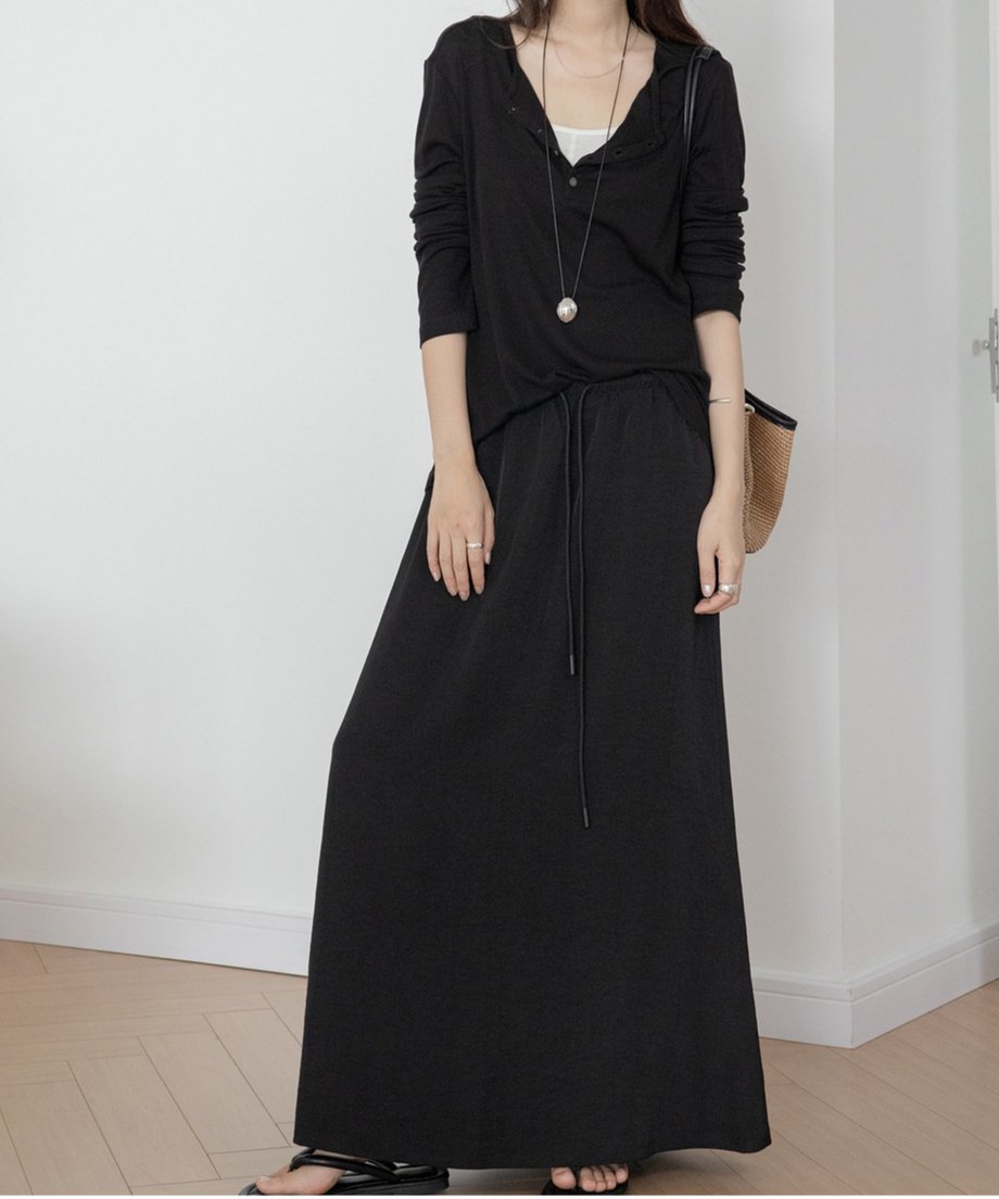 ブラックのドロストマキシスカートと黒い長袖カーディガンを着用した女性の全身。スカートはドローストリング仕様で、カゴバッグを肩にかけている。