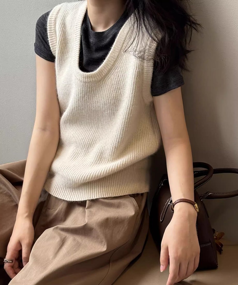 オフホワイトのUネックニットベストを黒いTシャツと茶色のワイドパンツにレイヤードして座っている女性の全身ショット。