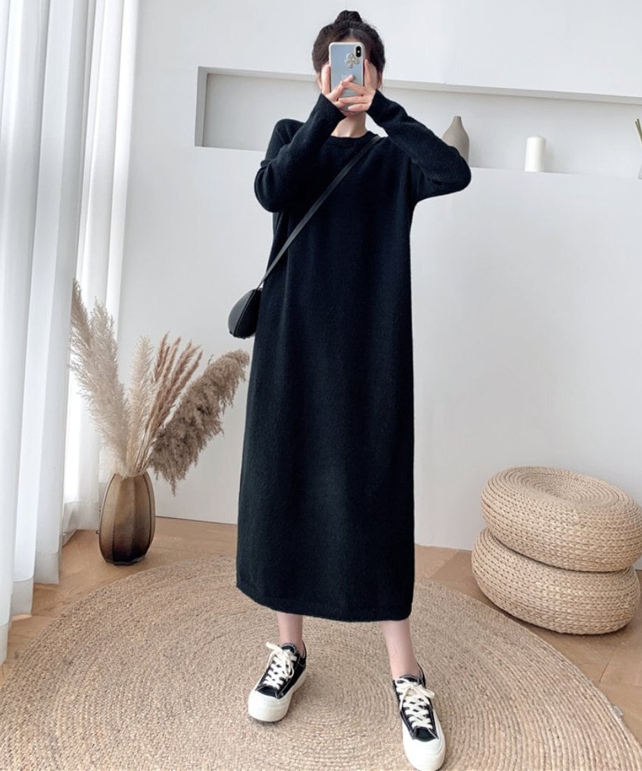 黒い長袖ニットワンピースを着用し、黒いショルダーバッグをかけた女性の全身。手にスマホを持っている。