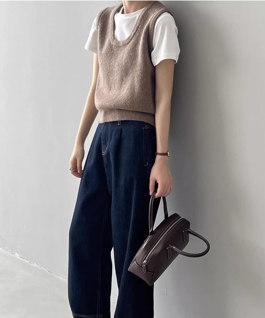ベージュのUネックニットベストを白Tシャツと濃い色のワイドデニムにレイヤードし、茶色のバッグを持っている女性の全身ショット。