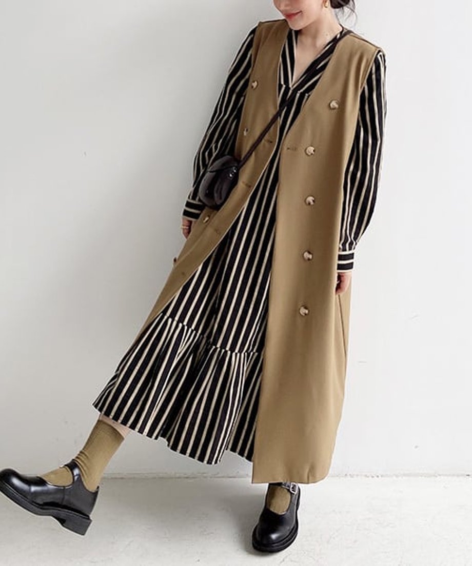 カーキのロングベストと黒白ストライプワンピースを着用した女性の斜め全身。黒のストラップシューズを履き、片足を上げている。