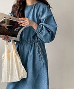 青のデニムドローストワンピースを着た女性のバストアップ。手に雑誌と白いバッグを持ち、ボリュームのある袖とウエストのドロストディテールが確認できる。