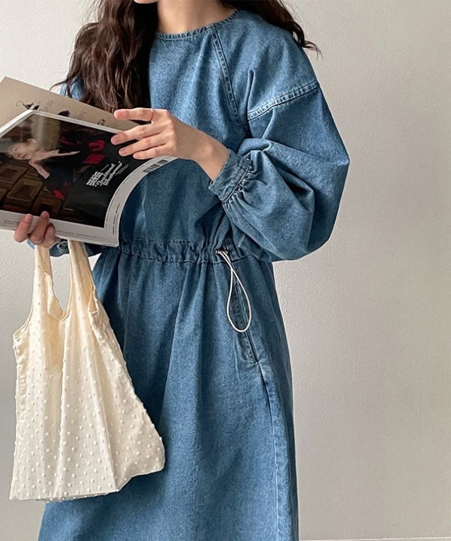 青のデニムドローストワンピースを着た女性のバストアップ。手に雑誌と白いバッグを持ち、ボリュームのある袖とウエストのドロストディテールが確認できる。