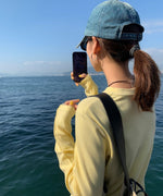 イエローのアウトシームロングTシャツを着て、海を背景にスマートフォンで写真を撮っている人物の後ろ姿。