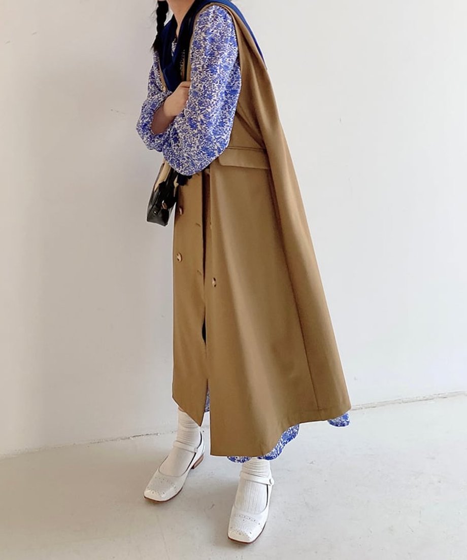 カーキのロングベストと青い花柄ワンピースを着用した女性の横向き全身。黒のバッグを持ち、白のソックスとローファーを着用。