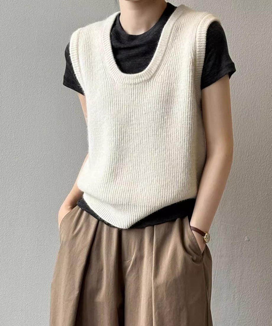 オフホワイトのUネックニットベストを黒いTシャツと茶色のワイドパンツにレイヤードして着用している女性のクローズアップ。
