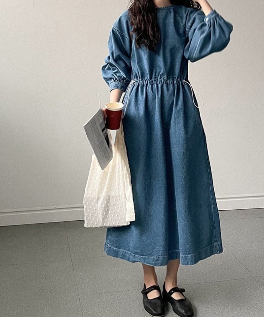 青のデニムのワンピースを着た女性の全身像。ウエストはドローストリングで絞られ、足元は黒いストラップシューズを着用。片手に紙コップと白いドット柄のバッグを持っている。
