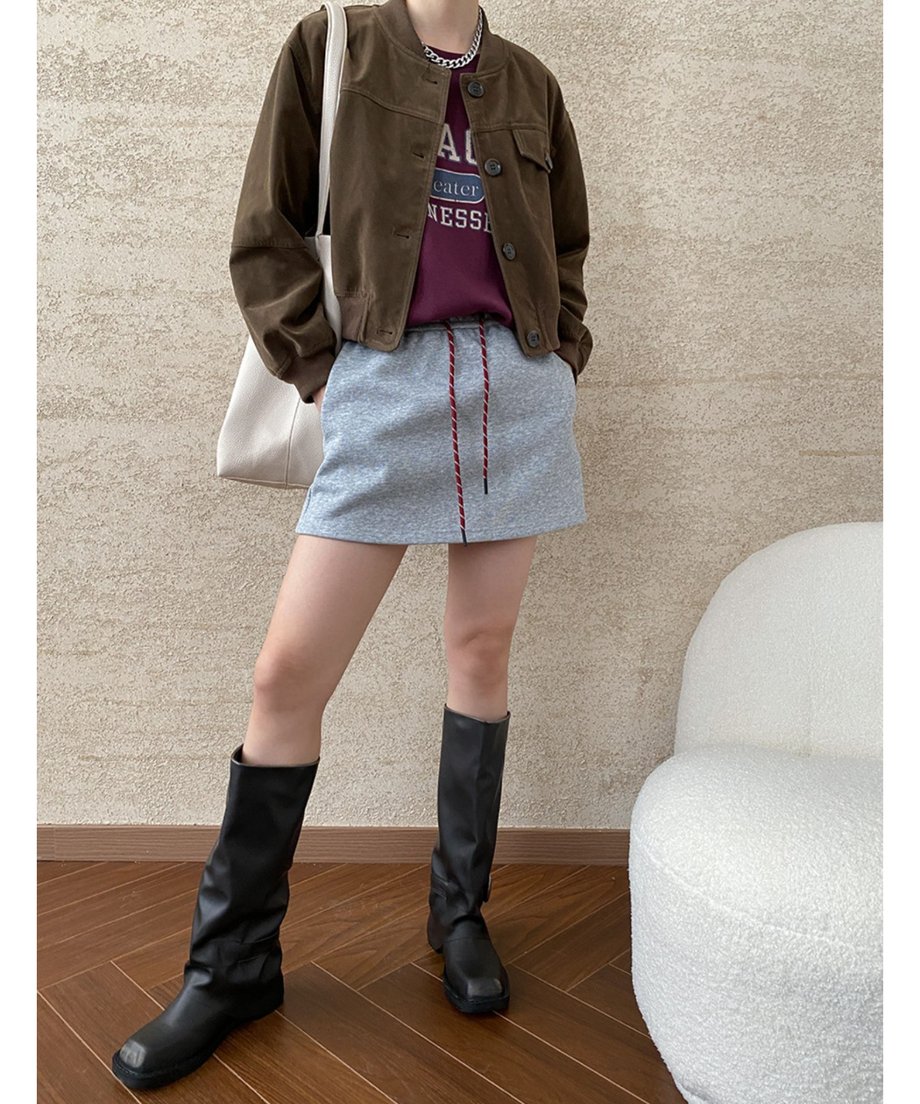 ブラウンのフェイクスエードジャケットを着用した女性の全身像。インナーはロゴTシャツ、ボトムスはグレーのミニスカート、足元は黒いロングブーツ。
