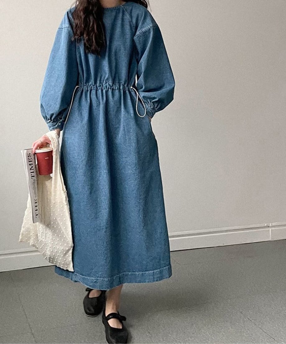 青のデニムドローストワンピースを着た女性の全身像。手に新聞と赤い紙コップ、白いバッグを持ち、ゆったりとしたシルエットで着用している。