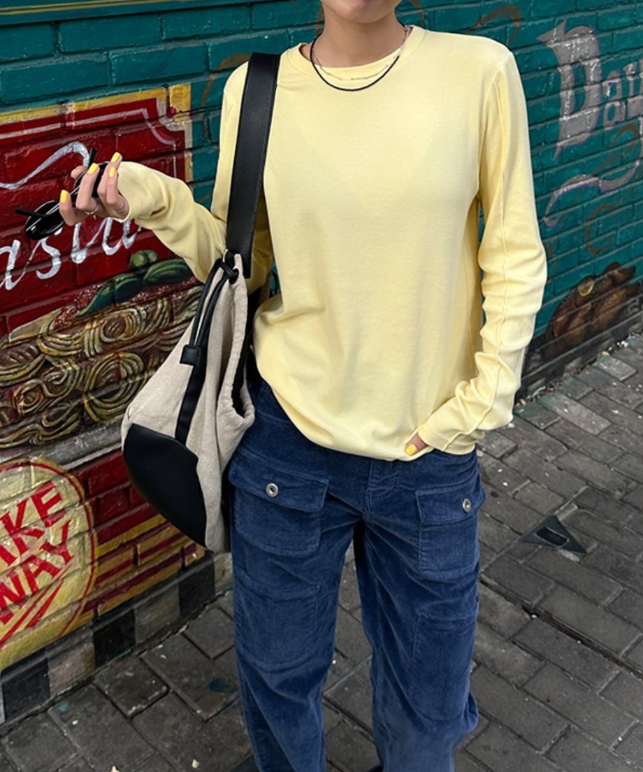 イエローのアウトシームロングTシャツを着て、ネイビーのコーデュロイパンツを着用した人物の全身。グラフィティアートの壁の前でポーズ。