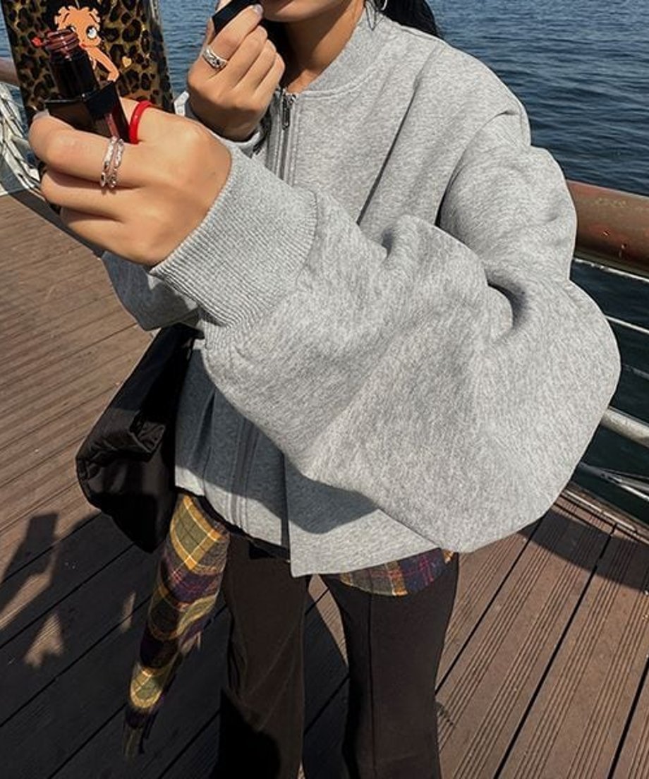 グレーのラージポケットブルゾンを着用し、屋外のウッドデッキでポーズをとる女性のバストアップ写真。黒いボトムスとチェック柄のストールが見える。