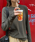 グレーのアウトシームロングTシャツを着て、ベージュのドローストリングパンツを着用した人物のバストアップ。赤い壁を背景に、手にはドリンクを持っている。