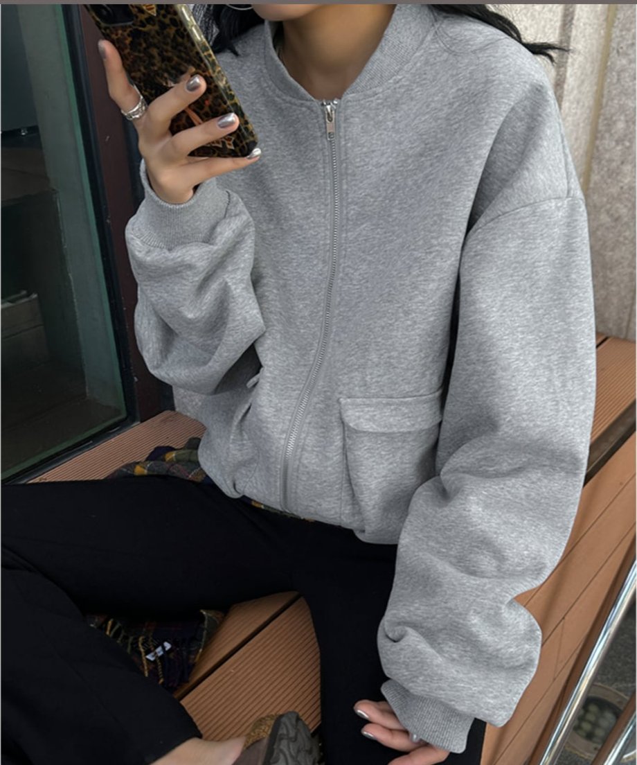グレーのラージポケットブルゾンを着用し、スマートフォンを持つ女性のバストアップ写真。大きなフラップポケットとオーバーサイズの袖が特徴。