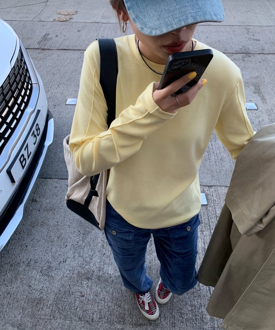 イエローのアウトシームロングTシャツとネイビーのコーデュロイカーゴパンツを着用し、デニムキャップを被った人物が携帯電話を見ている全身ショット。