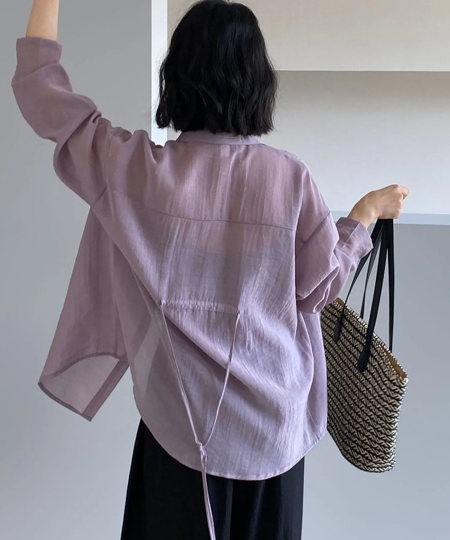 パープルのリボンシアーシャツを着用した女性の後ろ姿。手を上げ、柄物のカゴバッグを持っている。