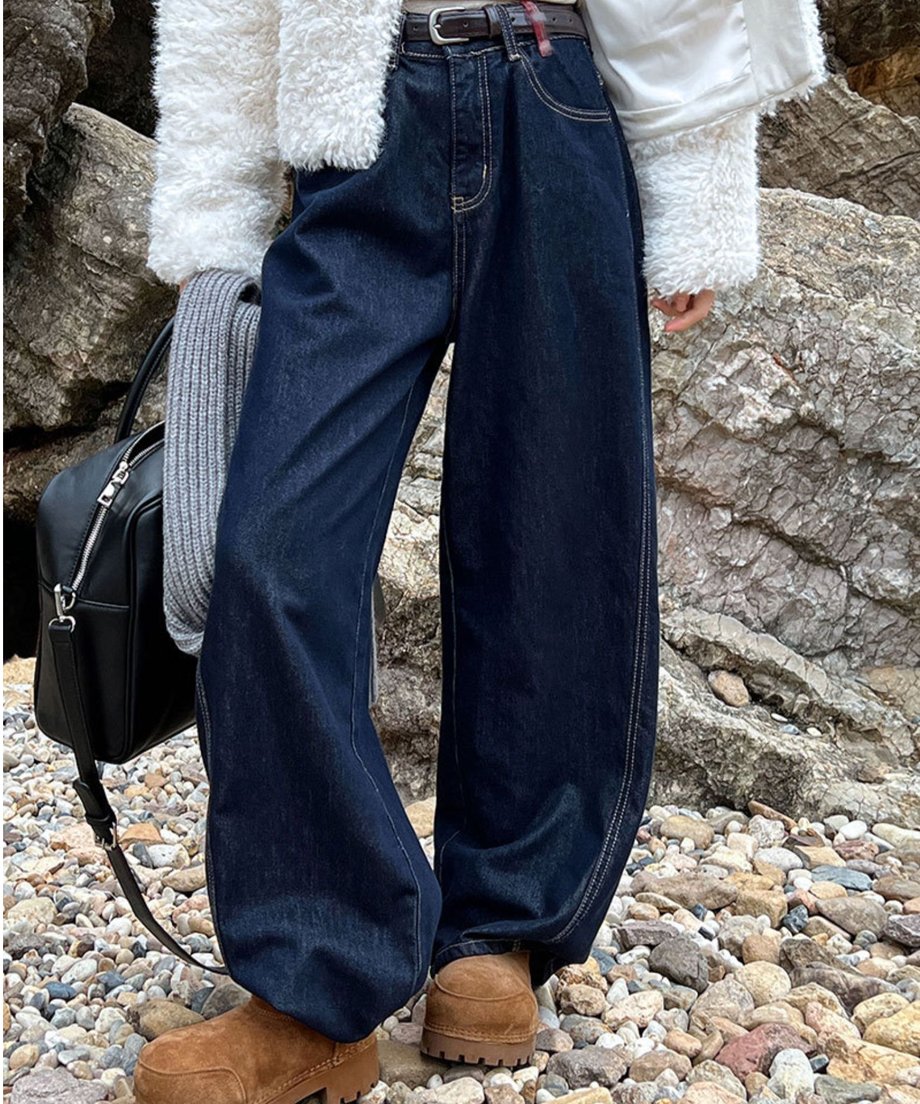 白いボアジャケットとダークブルーのワイドデニムパンツを着用した女性の全身。ベルトと茶色の厚底ブーツを合わせ、岩場で撮影されている。