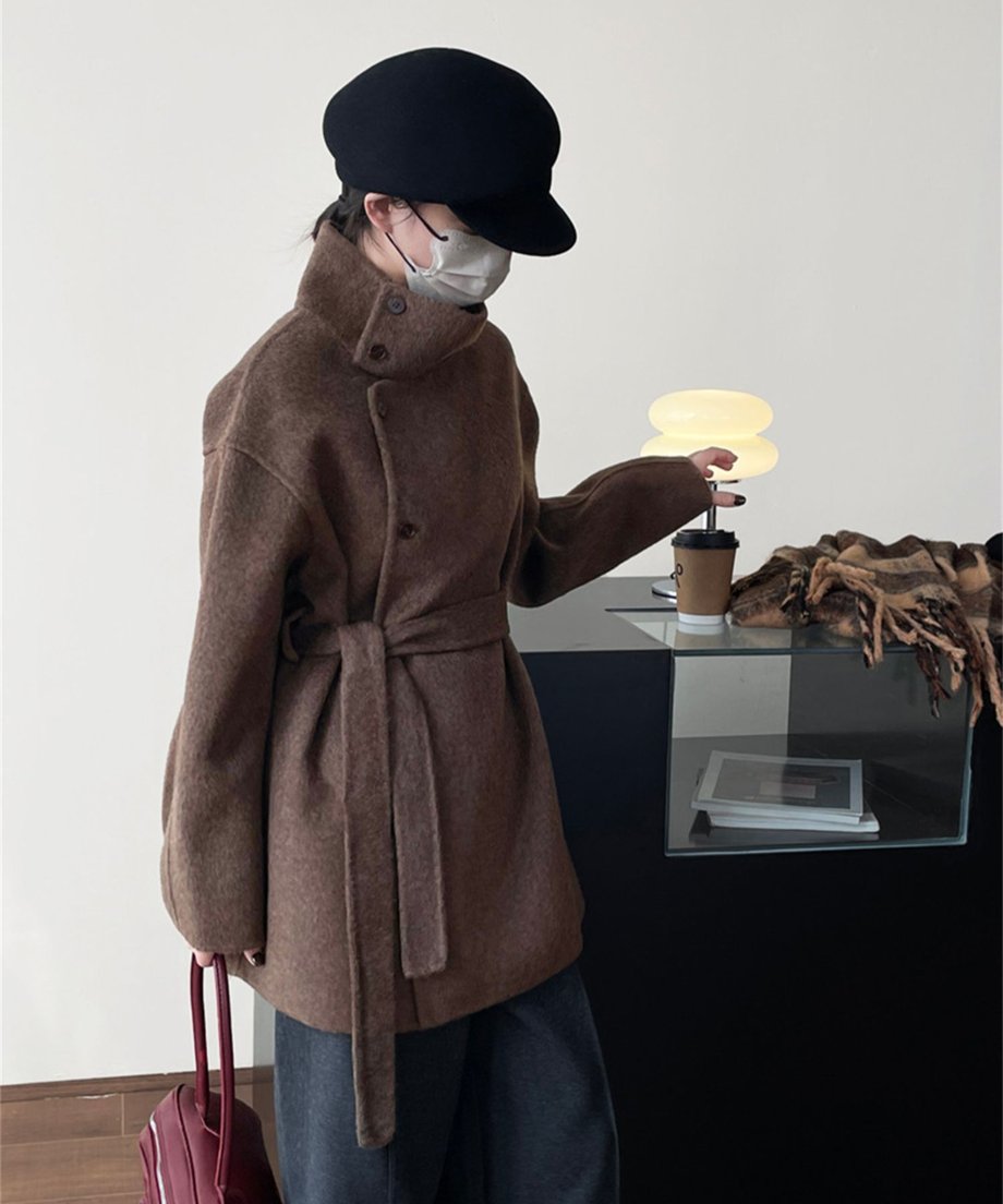 ブラウンのコートを着てカフェのテーブル横に立つ女性。襟を立てたデザインと横顔のシルエット、暖かみのある素材感。