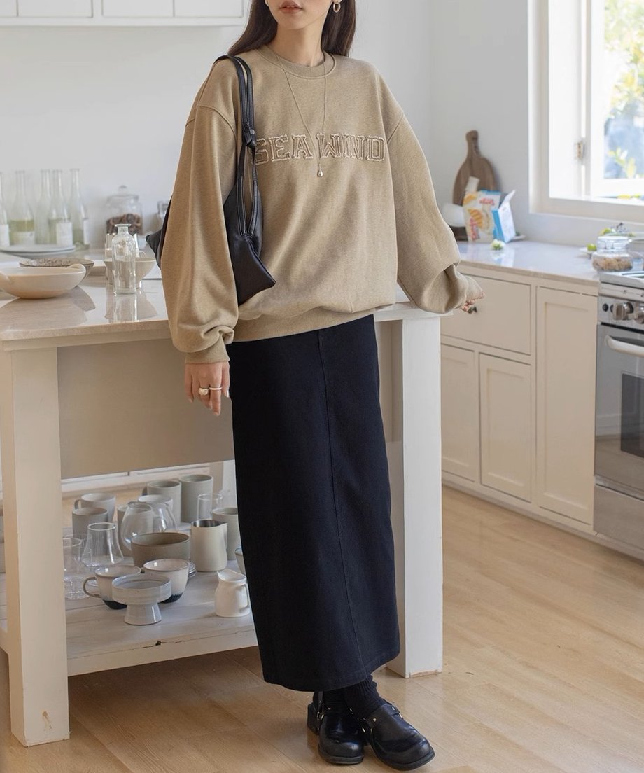 ベージュのロゴスウェットシャツを着た女性が黒のスカートを合わせて室内に立っている。