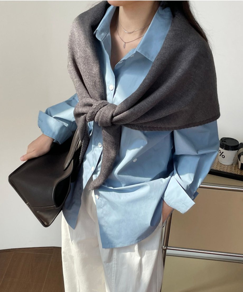 グレーのニットスカーフを、水色のシャツの上に前で結んで着用した女性のバストアップ。白のパンツと合わせたきれいめカジュアル。