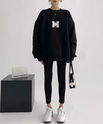 黒のオーバーサイズスウェットを着用している女性の全身ショット。白い大きな「M」のアップリケが見える。