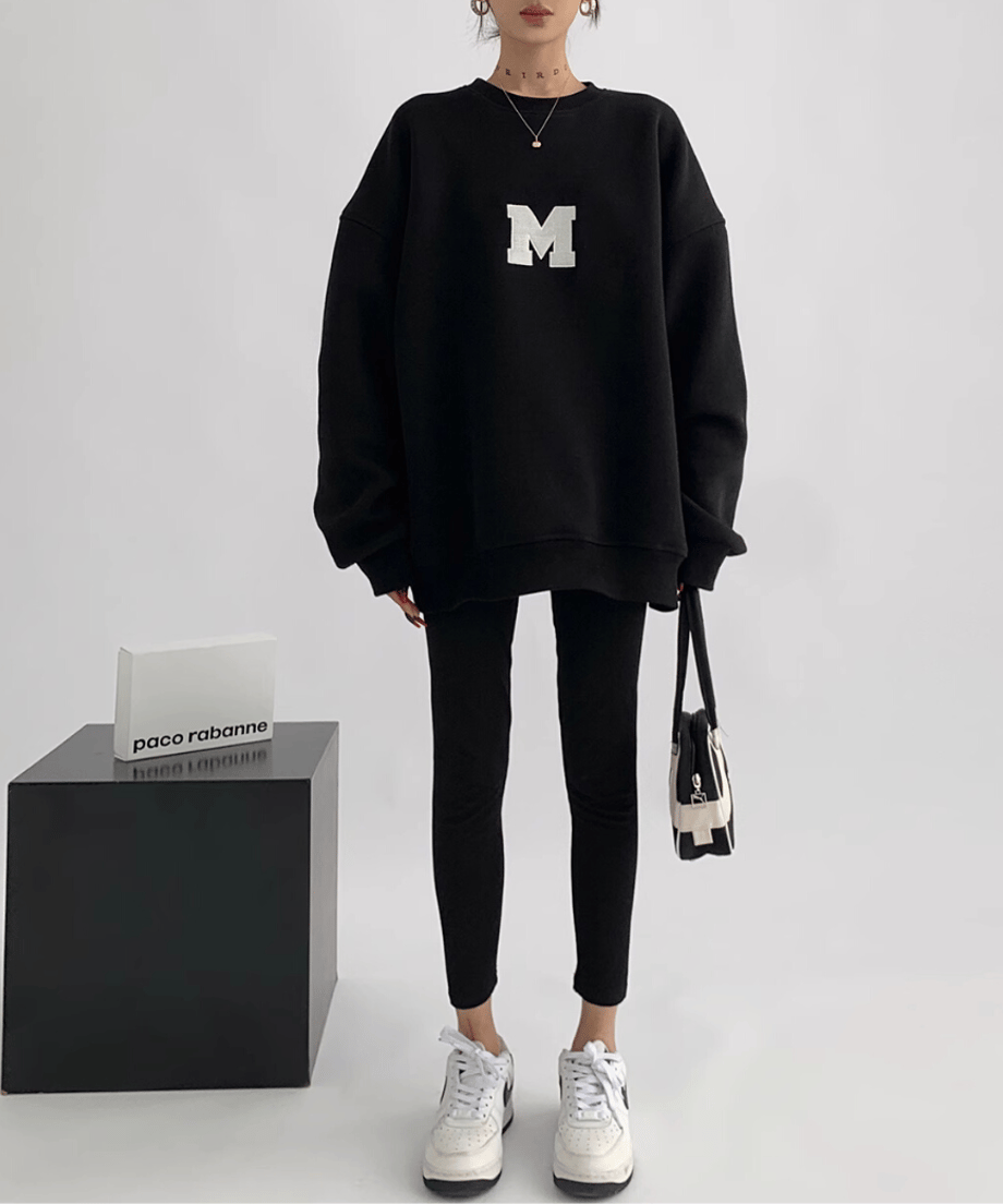 黒のオーバーサイズスウェットを着用している女性の全身ショット。白い大きな「M」のアップリケが見える。