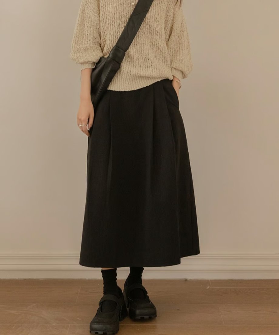 ベージュのニットと黒のプリーツスカートを着用した女性。クロスボディバッグとボリューム感のあるシューズがアクセント。