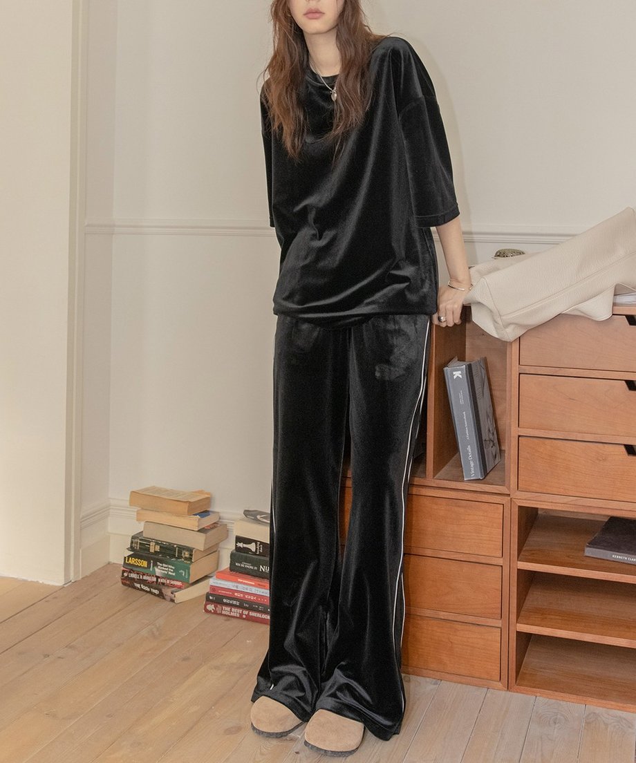 ブラックのベロアセットアップを着用した女性の全身。木製の棚の前に立ち、ゆったりとしたTシャツとワイドパンツでリラックスした雰囲気。